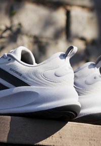 Chaussures de course blanches Nike Supernova Ease 2 avec des accents noirs sur la semelle, reposant sur une surface en pierre à l'extérieur sous la lumière naturelle.