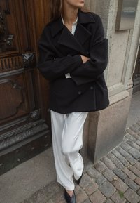 Musta villakangastakki, jossa on leveät läpät ja kaksirivinen nappikiinnitys, päällä valkoinen paita ja löysät vaaleanbeiget housut. Mustat teräväkärkiset kengät täydentävät asun.