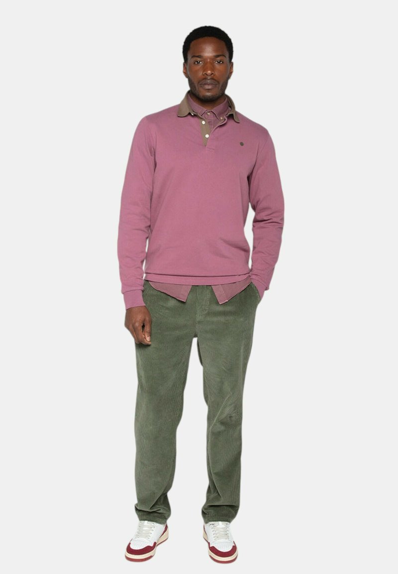Homme debout les mains dans les poches, portant une chemise longue rose, un pantalon en velours côtelé vert olive et des baskets blanches avec des accents rouges.