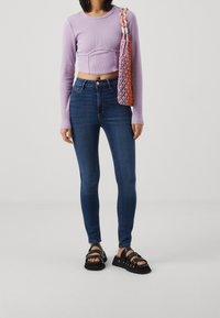 Lila långärmad kort topp, blå skinnyjeans och svarta kraftiga sandaler med flera remmar. En flerfärgad nätväska hänger över axeln.