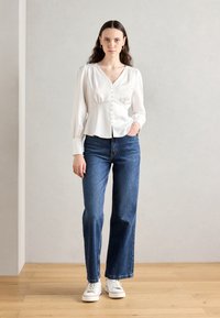 Blouse blanche à manches longues avec boutons sur le devant, décolleté en V et épaules froncées, associée à un jean bleu à jambes larges et des baskets blanches.