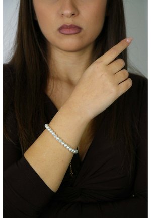 Biała bransoletka z pereł o gładkim, błyszczącym wykończeniu, noszona na nadgarstku. Osoba wskazuje drugą ręką.