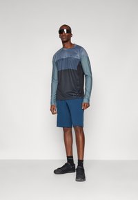 Long-sleeve shirt in blue and gray with geometric color blocks; paired with dark blue shorts and black athletic shoes. Reflective sunglasses worn.