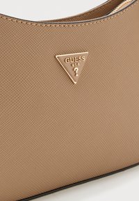 Beige handtassectie met textuur en een gouden driehoekig Guess USA-logo met daarin een vraagteken.