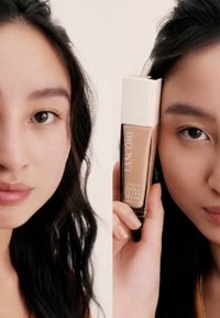 Bouteille de fond de teint dans un récipient en verre transparent avec un capuchon blanc, tenue à côté du visage d'un mannequin ; texture lisse, couleur beige claire, étiquetée "Lancome."