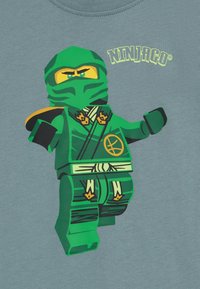 Žalias LEGO Ninjago personažas ant šviesiai mėlynos marškinėlių. Rodo detalią veido išraišką, nindzios aprangą ir priedus.
