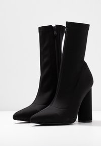 Bottes à talons hauts noires avec une tige en tissu lisse et extensible, un bout pointu et une fermeture éclair à l'intérieur de la cheville.