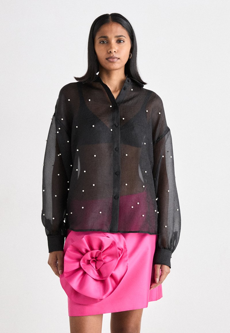 Schwarze transparente Knopf-Bluse, verziert mit weißen Perlen, kombiniert mit einem leuchtend pinken Rock, der ein großes, gefranstes Blumendetail aufweist.