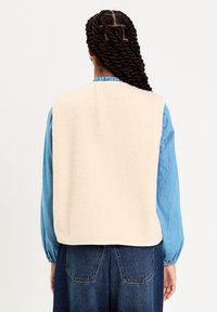 Gilet en polaire de couleur crème, avec un col montant et des coutures plates, porté sur une chemise bleue avec des poignets froncés et un jean en denim.