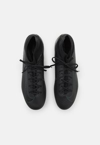 Des crampons de football noirs avec un motif en chevrons texturé, présentant des lacets ton sur ton et un subtil swoosh bleu de Nike. Design léger.