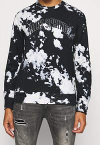 Sudadera de tie-dye en blanco y negro con el texto "PALM SPRINGS", cuello redondo y puños acanalados. Combinada con jeans grises desgastados.