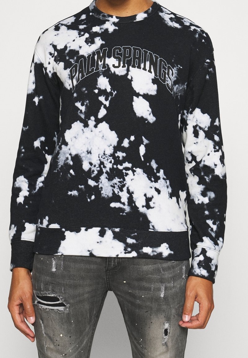 Sudadera de tie-dye en blanco y negro con el texto "PALM SPRINGS", cuello redondo y puños acanalados. Combinada con jeans grises desgastados.