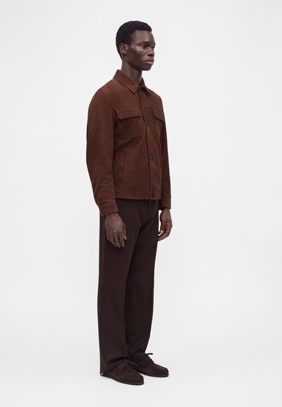 Homme debout portant une veste en daim marron, un pantalon marron assorti et des chaussures marron foncé à lacets sur un fond blanc uni.