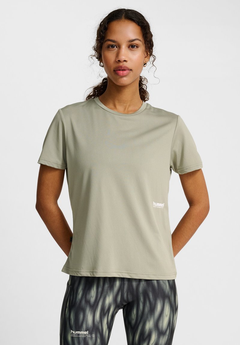 Jeune femme portant un t-shirt de sport gris clair à manches courtes et des leggings noirs et gris à motifs, debout les mains derrière le dos sur fond blanc.