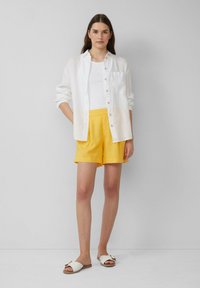 Chemise en lin blanche à boutons sur un débardeur blanc, associée à un short jaune et des sandales blanches avec un accent doré. Design minimaliste.
