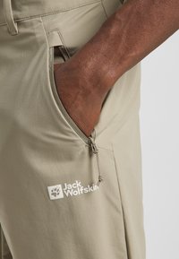 Beige Wanderhose mit einer Reißverschlusstasche. Das Material wirkt leicht und glatt. Auf der Vorderseite befindet sich ein weißes Logo.