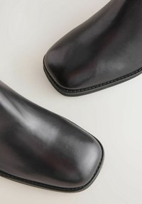 Gros plan sur deux bottes en cuir noir à bouts carrés sur un fond clair uni.