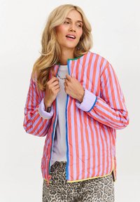 Kobieta z falującymi blond włosami, ubrana w kurtkę w czerwono-fioletowe paski nad białą koszulą i spodniami w panterkę, patrzy na bok.