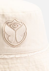 Chapeau seau beige en tissu texturé, présentant un emblème brodé avec un œil stylisé et des ailes, ainsi qu'un détail de bord effiloché.