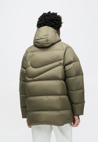 Olivo zelena podložena jakna s kapuco, s prešito zasnovo in izrazitim logotipom Nike na hrbtu. Teksturirana, vodoodporna tkanina.