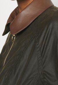 Gros plan sur une veste vert foncé avec un col en cuir marron et un détail de fermeture éclair dorée sur le haut du torse d'une personne.