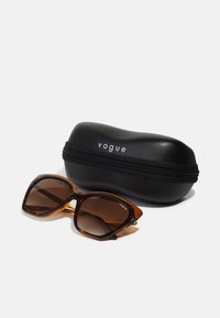 Lunettes de soleil écaille de tortue avec verres marron, design angulaire et accent doré sur les branches, accompagnées d'une housse zippée noire étiquetée "vogue".