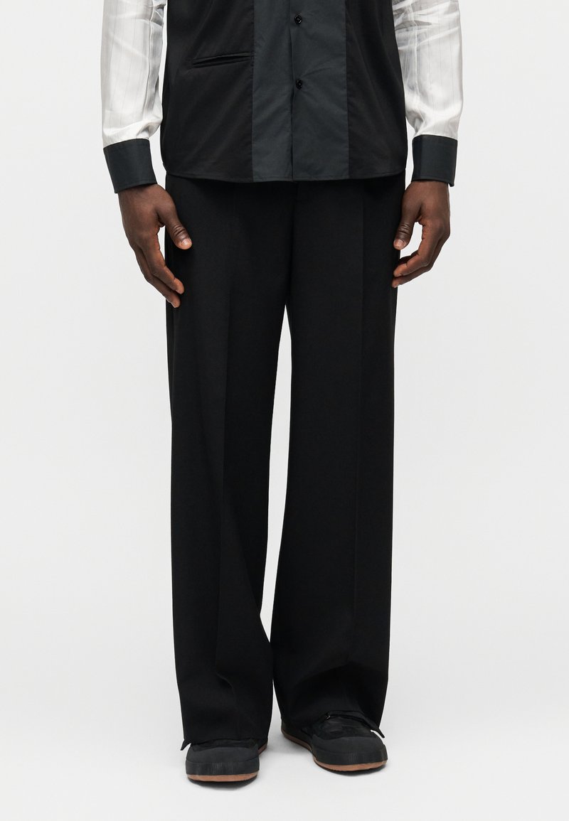 Pantalon large noir avec une texture lisse, présentant une coupe classique. Le mannequin porte des chaussures foncées, complétant l'ensemble du look.