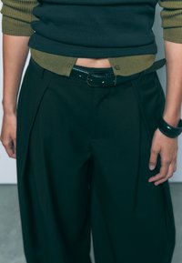 Široke črne hlače s gubami, v kombinaciji s temnim zgornjim delom in zelenim rebrastim pasom. Obleko dopolnjuje sijoč črn pas.