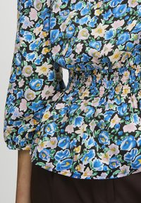 Blusa floreale con sfondo nero, fiori blu, rosa e gialli, vita arricciata e maniche a palloncino per un volume extra.