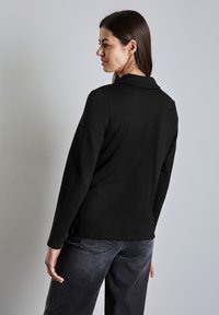 Svart långärmad skjorta med krage, texturerad tyg och en slät finish, bärs med grå jeans. Utsikt bakifrån som visar en minimal design.