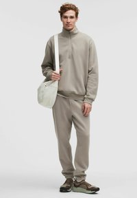 Jonge man in een beige trainingspak met een half-rits pullover, bruin en beige sneakers, die een crèmekleurige schoudertas vasthoudt tegen een effen achtergrond.