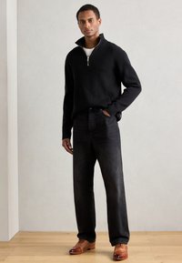 Pullover nero a coste con colletto mezza zip, abbinato a jeans scuri e scarpe in pelle intrecciata marroni su un pavimento in legno chiaro.