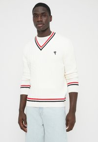 Balta megzta megztinis su V formos iškirpte, puoštu pynimo raštais ir juodomis, raudonomis bei baltomis juostelėmis prie kaklo apykaklės ir rankogalių. Juodas emblemą ant krūtinės.