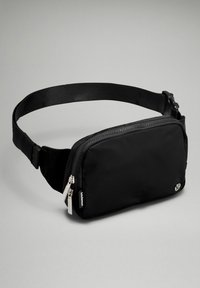 Bolso de cintura de tela negra con forma rectangular, cierre con cremallera, correa ajustable y acentos de hardware plateados. Etiquetado con un logotipo.