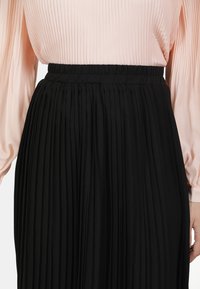 Vrouw in een geplooide zwarte rok met een elastische taille en een lichtroze blouse met lange mouwen die erin is gestoken.