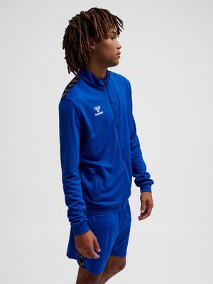 Jonge man met dreadlocks die een blauwe hummel sportjas en bijpassende blauwe korte broek draagt, staat en kijkt naar rechts.
