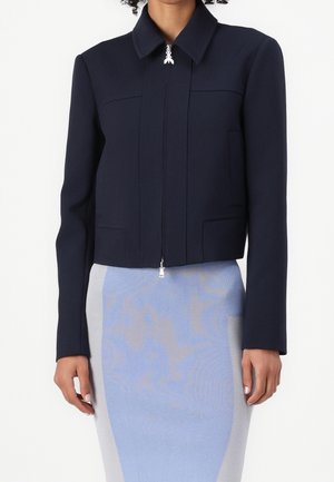 Kvinde iført en tætsiddende, mørkeblå lynlåsjakke over et lyseblåt og gråt pencilskørt, stående mod en ensfarvet baggrund.