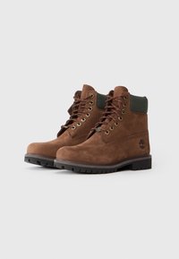 Botas de tobillo de ante marrón con un cuello de tela verde, punta redonda, ojales de metal y suela de goma negra y resistente. Presenta un bordado del logo en el lateral.