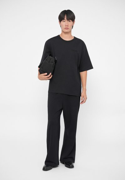 Camiseta negra de gran tamaño con mangas cortas, combinada con pantalones negros de pierna ancha. El modelo sostiene un pequeño bolso negro. Diseño simple y monocromático.