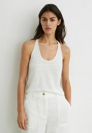 Femeie cu păr lung și întunecat, purtând un top alb fără mâneci și pantaloni albi cu talie înaltă, pe un fundal alb simplu.