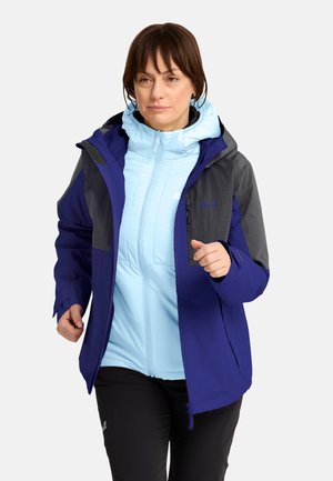 Frau trägt eine blaue und graue Outdoorjacke über einem hellblauen Zip-Hoodie und steht vor einem schlichten weißen Hintergrund.