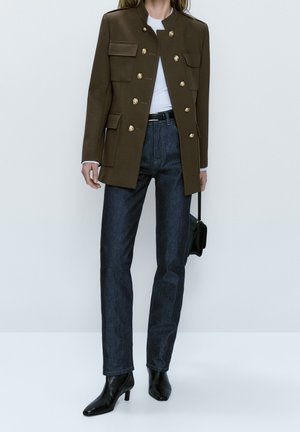 Femme portant une veste militaire vert olive avec des boutons dorés, un jean bleu foncé, des bottines noires et tenant un sac à main noir.