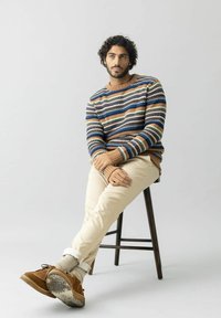 Vīrietis ar tumšiem cirpotiem matiem sēž uz koka krēsla, ģērbtā svītrainā džemperī, bežās biksēs, pelēkās zeķēs un brūnās zamša kurpēs.