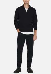 Sort strikket trøje med en halv-zip krave, stylet over en hvid skjorte, parret med sorte bukser og sorte sko, alt sammen i en neutral indstilling.