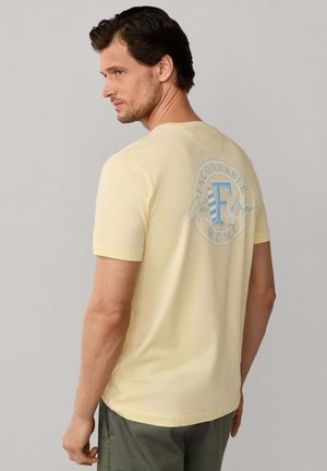 Homme portant un t-shirt jaune pâle à manches courtes avec un logo circulaire bleu clair et un texte au dos, associé à un pantalon gris.