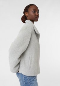 Cappotto in lana grigio chiaro, dalla texture lavorata, con colletto a zig zag, due tasche laterali e una vestibilità leggermente cropped, abbinato a jeans in denim.