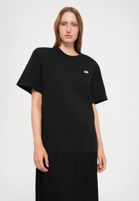 Young woman with straight red hair wears an oversized black T-shirt with a small embroidered emblem on the left chest against a white background.
