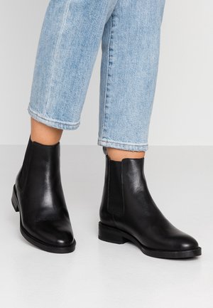 Bottes en cuir noir montant à la cheville portées avec un jean bleu clair à revers, présentées sur un sol blanc avec un fond clair uni.