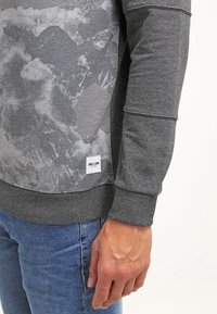 Personne portant un sweat gris à motifs avec poignets et ourlet côtelés, un jean bleu et se tenant devant un fond blanc.