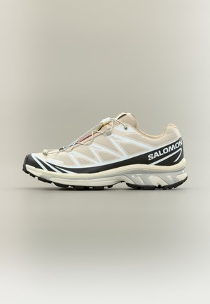 Chaussure de sport légère avec une tige en mesh beige, des accents blancs, et une semelle en caoutchouc noire. Dispose d'un système de laçage rapide et d'un talon texturé.